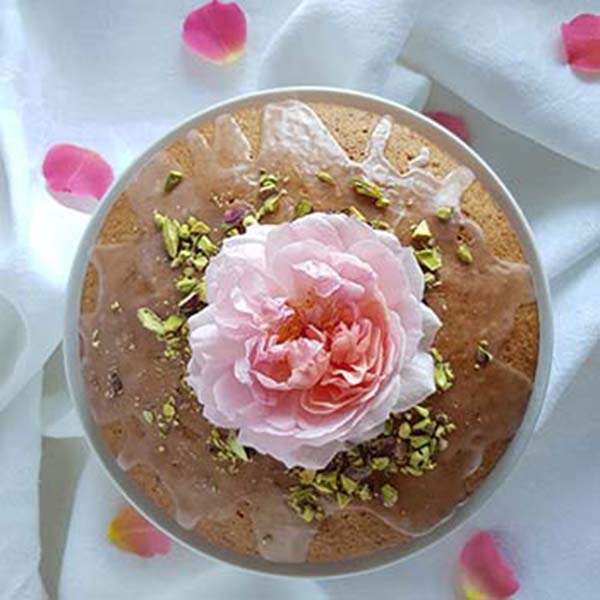 Sciogliere il burro e unirlo allo zucchero. Aggiungere le uova, uno alla volta. Unire il succo e la scorza di mezza arancia. Tritare finemente i pistacchi e le mandorle, unirli all’impasto insieme alla farina, alla fecola e al lievito setacciati. Cuocere in una teglia imburrata e infarinata di 21 cm per 40 minuti a 180 °C. Preparare la glassa: stemperare lo zucchero a velo con l’acqua alle rose e l’acqua naturale. Io ho aggiunto una goccia di colorante alimentare rosa per dare un po’ di colore alla glassa. Versare la glassa sulla torta e decorare subito con pistacchi tritati e petali di rosa.